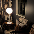 A modern spherical table lamp with a glowing white globe shade and white base, placed on a dark wooden side table next to a striped armchair in a dimly lit room.