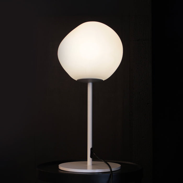 A modern table lamp with a glowing white organic-shaped globe shade, a slim white stem, and a round white base, set against a dark background.