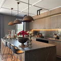 A modern kitchen interior with a large island, dark gray marble countertop, light wood cabinetry, three bronze pendant lights, and a red poinsettia arrangement on the counter.